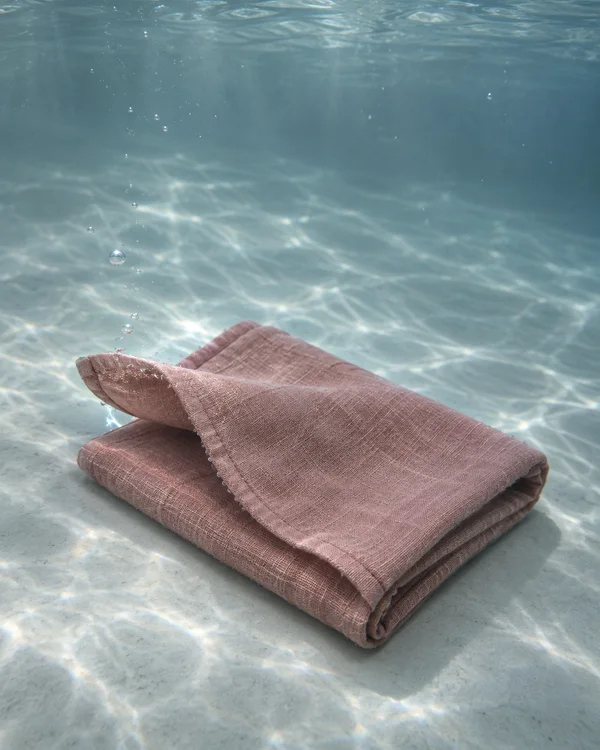 A dusty-rose linen dishcloth lies folded on a pale tank floor underwater, with one hem lifted into a broad curved flap; caustic light and bubbles are visible…