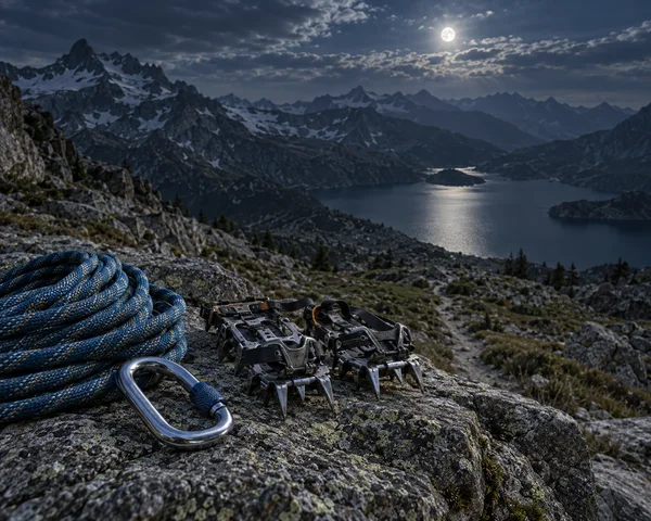 A coiled blue climbing rope, a metal carabiner, and two crampons sit on a rocky ledge above a mountain lake.