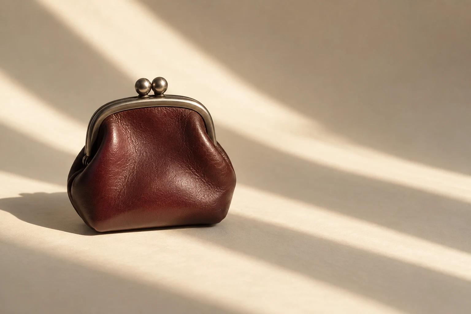 A closed oxblood leather coin purse with a rounded nickel kiss-lock frame and two spherical knobs sits left of centre on an ivory surface crossed by diagonal…