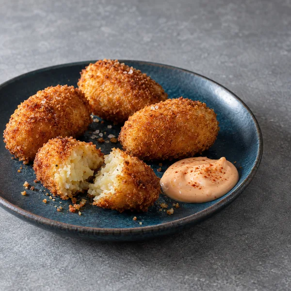 Three whole golden-brown potato croquettes and two pieces of a split croquette sit on a dark petrol-blue plate with pale paprika-speckled aioli, on a grey…