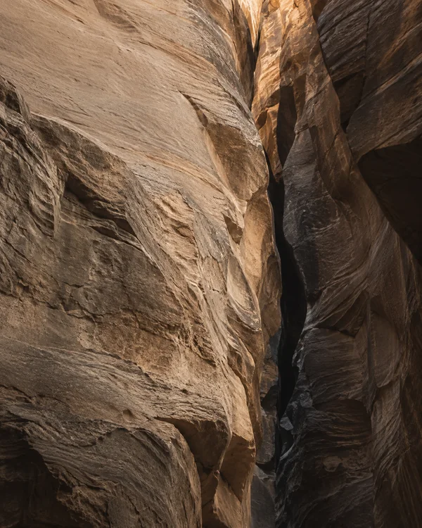 A tight upward-looking view between massive sandstone canyon walls, with a pale, striated rock face on the left and a dark narrow fissure just right of center.