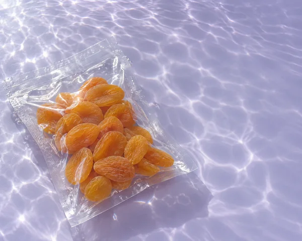 A clear heat-sealed pouch of golden-orange dried apricots lies diagonally on a lavender-toned surface beneath rippling water, with bright caustic patterns and…