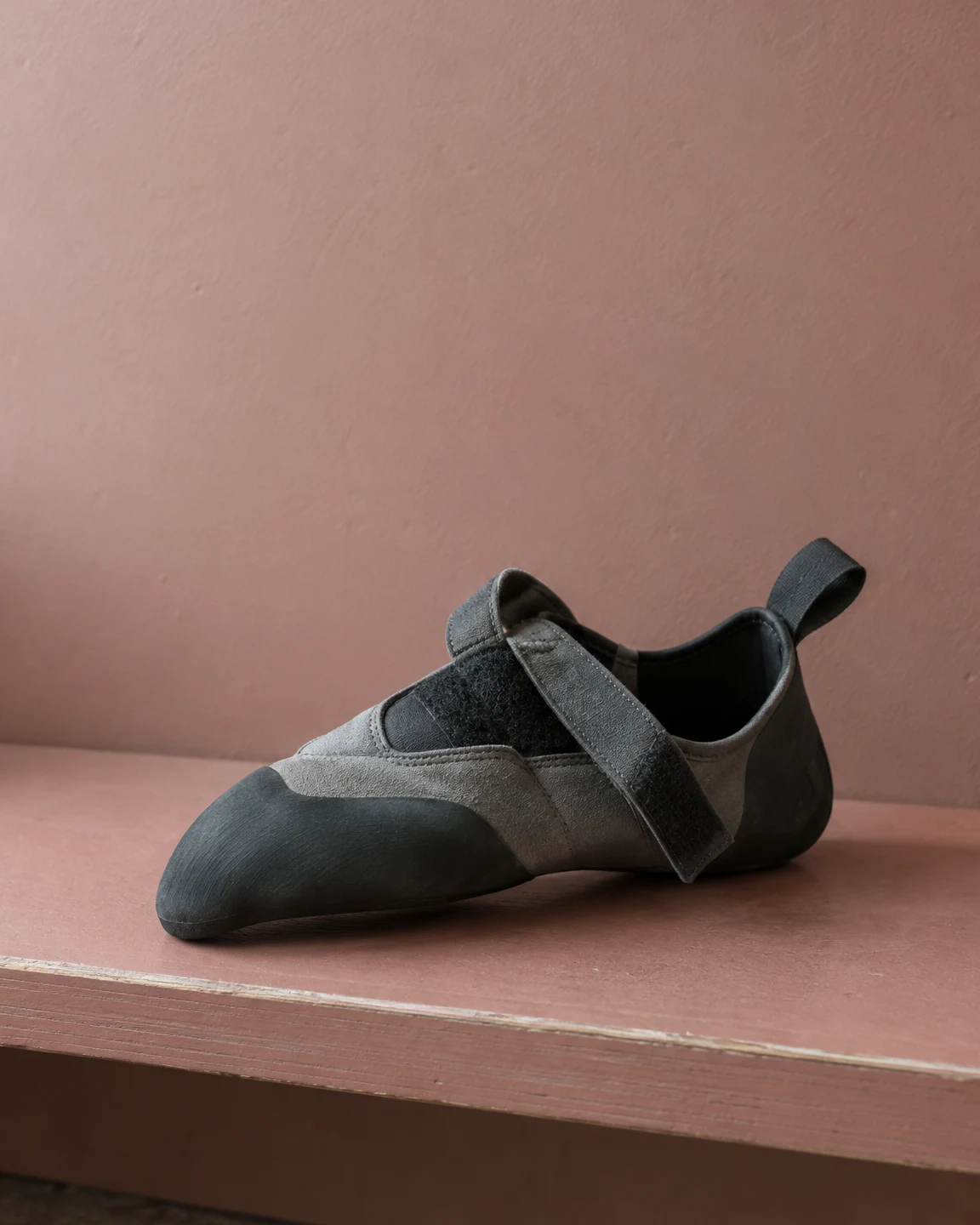 A slate-grey climbing shoe rests on its side on a dusty-rose plywood bench, its black rubber toe pointing toward the lower left and a loose strap draped across…