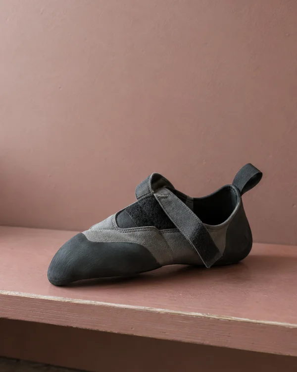A slate-grey climbing shoe rests on its side on a dusty-rose plywood bench, its black rubber toe pointing toward the lower left and a loose strap draped across…