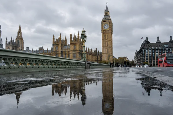 Low-angle view from a wet Westminster Bridge pavement toward Elizabeth Tower and the Palace of Westminster, with a broad puddle reflecting the tower, bridge…