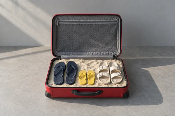 An open red hard-shell suitcase sits on a grey surface, its lower compartment filled with pale sand and holding navy flip-flops, off-white buckled sandals and…