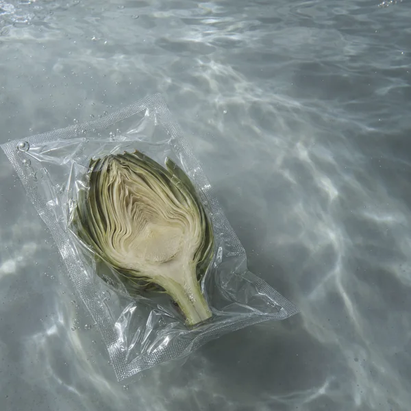 A lengthwise artichoke half sits inside a clear sealed pouch on a pale gray tank floor, with rippling water reflections and small bubbles around it.