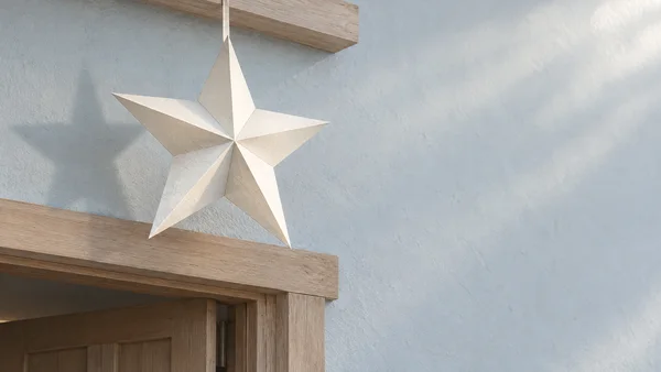 A large ivory folded-paper star hangs from a narrow ribbon beneath a light wooden lintel above a cropped wooden doorway, against a pale blue-gray plaster wall…