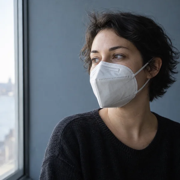 A short-haired adult wearing a plain white folded respirator looks toward a bright window, framed from the chest up against a blue-grey wall.