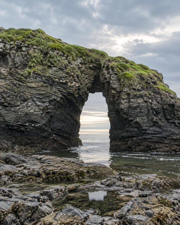A dark, layered sea arch topped with bright green grass frames a pale strip of ocean beneath a cloudy sky.