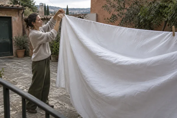 A woman in a knitted sweater fastens a large white sheet to a clothesline in a stone-paved courtyard, viewed from above with a dark railing in the foreground.