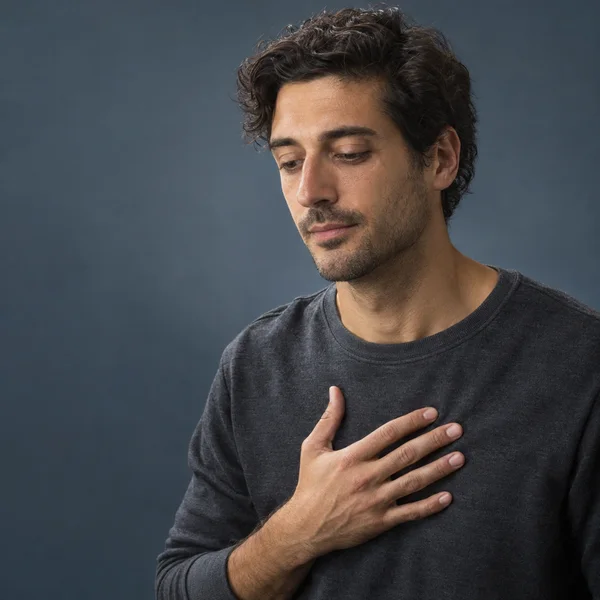 A man in a charcoal crewneck looks downward with one hand resting on his chest, photographed against a muted blue-gray background with open space to the left.