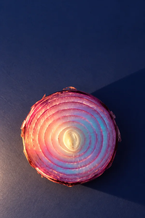 A cross-section of a red onion rests cut-side up on a textured dark indigo surface, its concentric rings glowing pink, coral, cream and cyan.