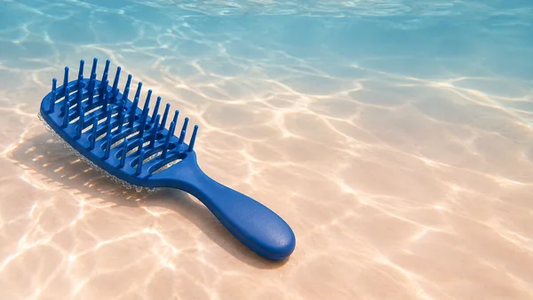 A cobalt-blue vented detangling brush rests diagonally on a pale peach surface, with its teeth facing upward, beneath rippling underwater light.
