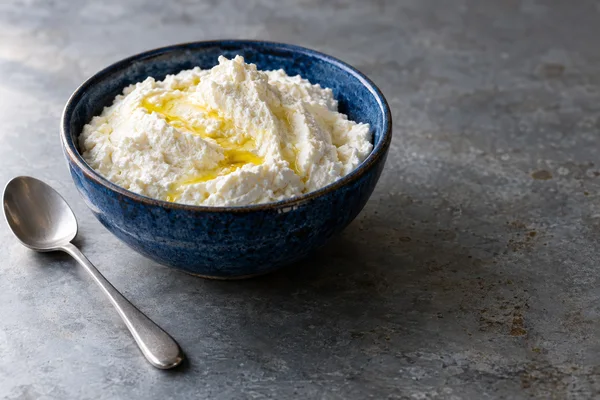 A cobalt-blue stoneware bowl filled with ivory ricotta and golden olive oil sits on a weathered grey tabletop, with a stainless-steel spoon beside it and open…