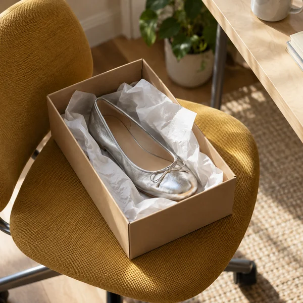 A silver metallic ballet flat rests in an open kraft shoebox lined with crumpled white tissue on a mustard swivel chair beside a pale wood desk, with a plant…