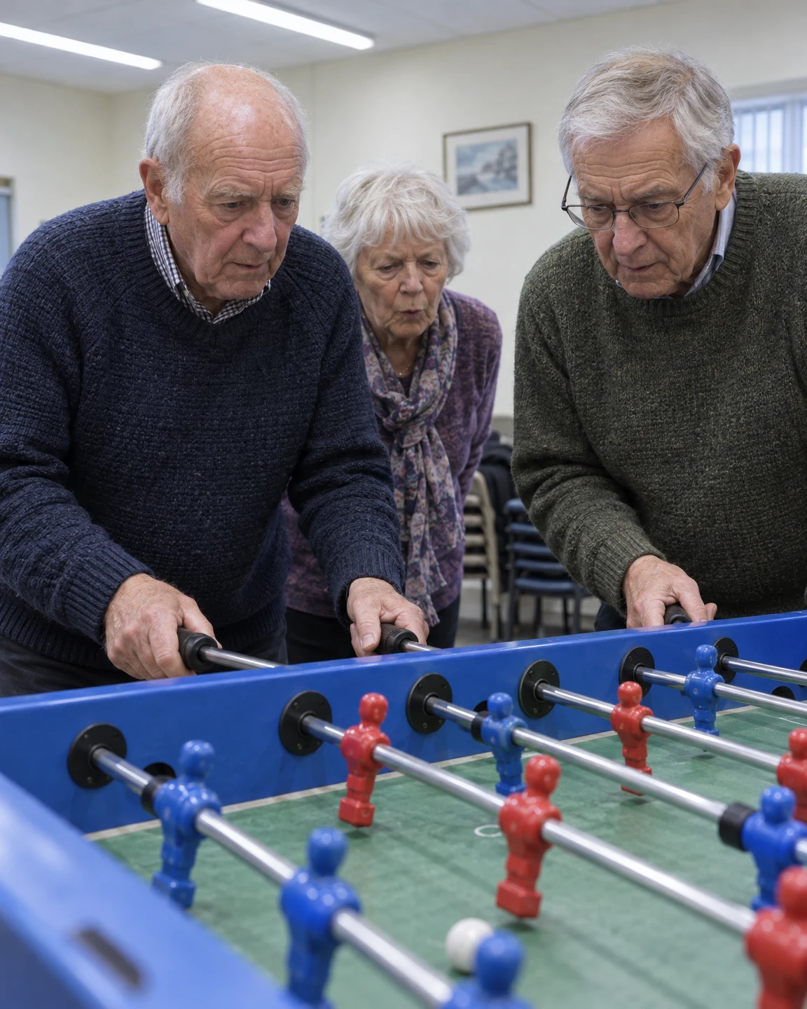Three older adults in knitwear lean over a blue foosball table in a pale-walled room.