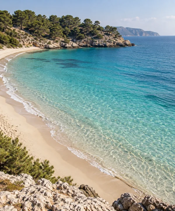 A curved pale-sand beach borders clear aquamarine shallows in a sheltered Mediterranean bay, with a pine-covered limestone headland and distant blue coastline…