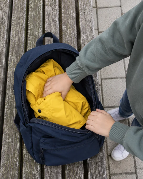 Overhead view of a child pushing a bright yellow rain jacket into an open navy backpack resting on a weathered wooden bench beside pale square paving; the…