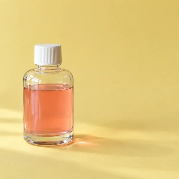 A clear cylindrical bottle with a ribbed white screw cap and coral-pink liquid stands on a butter-yellow surface, with broad window reflections on the glass…