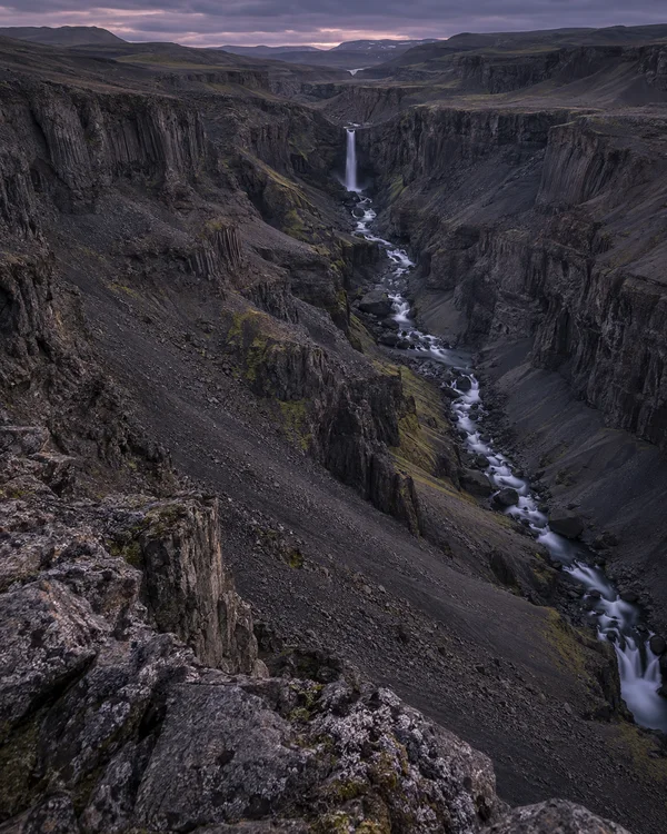 A steep view into a rugged, dark-rock gorge at dusk, with a narrow waterfall feeding a silky stream that winds between the cliffs beneath a muted purple sky.