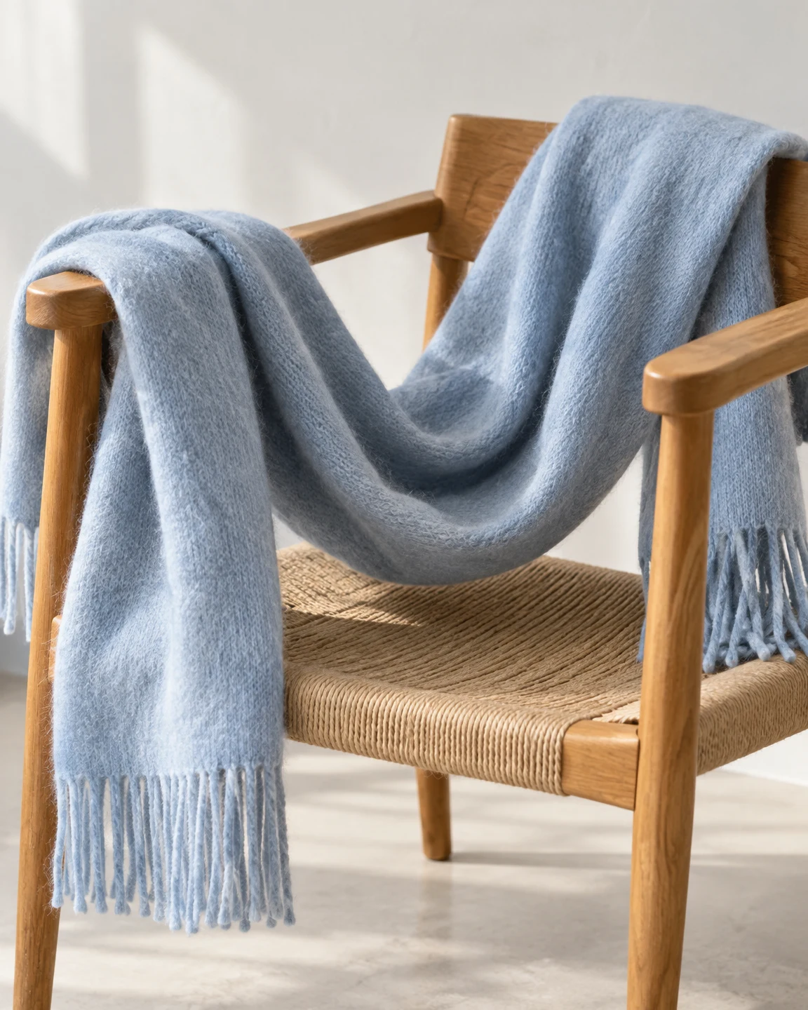 A powder-blue fringed scarf drapes over the arms of a honey-brown wooden armchair, sagging in a deep U above its woven seat against a pale studio wall and…