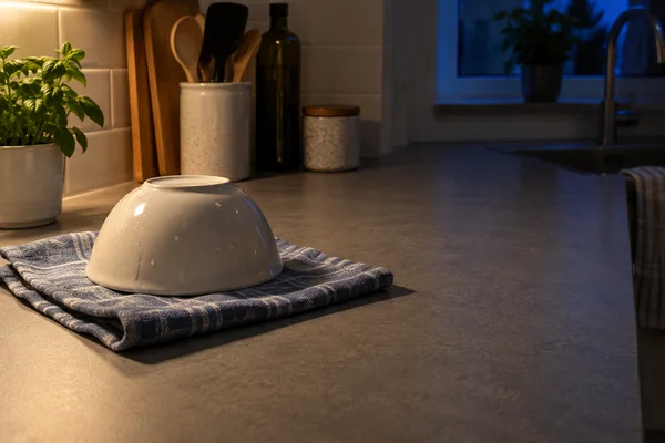 An ivory ceramic bowl sits upside down on a folded blue patterned tea towel on a grey kitchen counter, with warm light from the left and a cool blue window and…