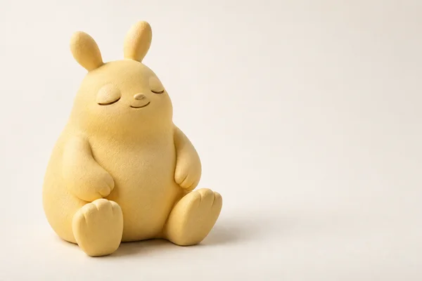 A butter-yellow, pear-shaped creature figurine sits with its broad feet extended on a warm ivory surface.