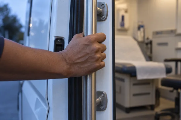 A medium-brown hand grips a vertical brushed-metal rail beside the open white doorway of a vehicle; a paper-covered examination couch and clinical equipment…