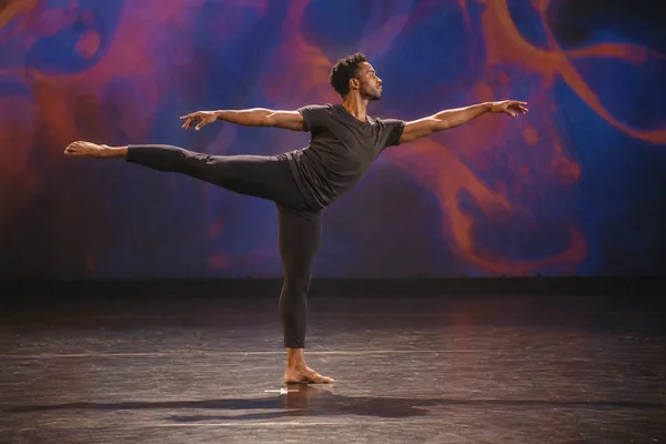 A Black male dancer balances barefoot on one leg on a theater stage, with the other leg extended horizontally and both arms outstretched; colorful abstract…