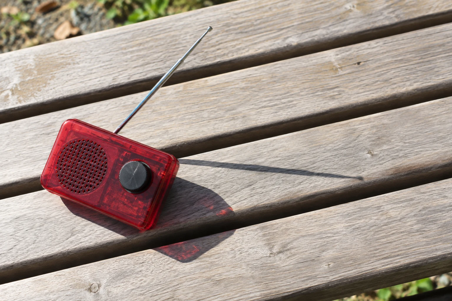 A translucent red pocket radio with a silver telescopic antenna rests at an angle on a sunlit pale wooden bench, casting a dark shadow and a fine antenna…
