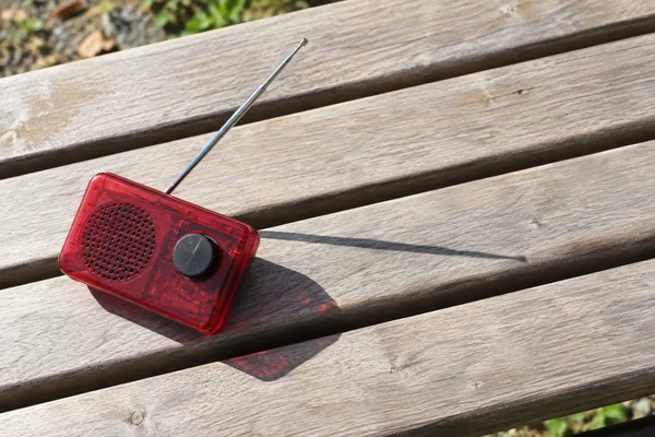 A translucent red pocket radio with a silver telescopic antenna rests at an angle on a sunlit pale wooden bench, casting a dark shadow and a fine antenna…