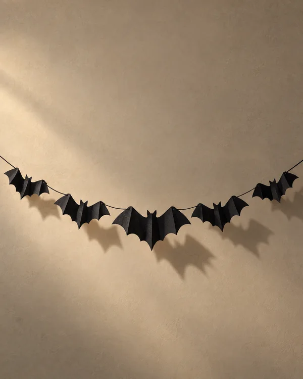 A curved garland of five folded black paper bats hangs across a warm beige textured backdrop, casting long shadows to the right.