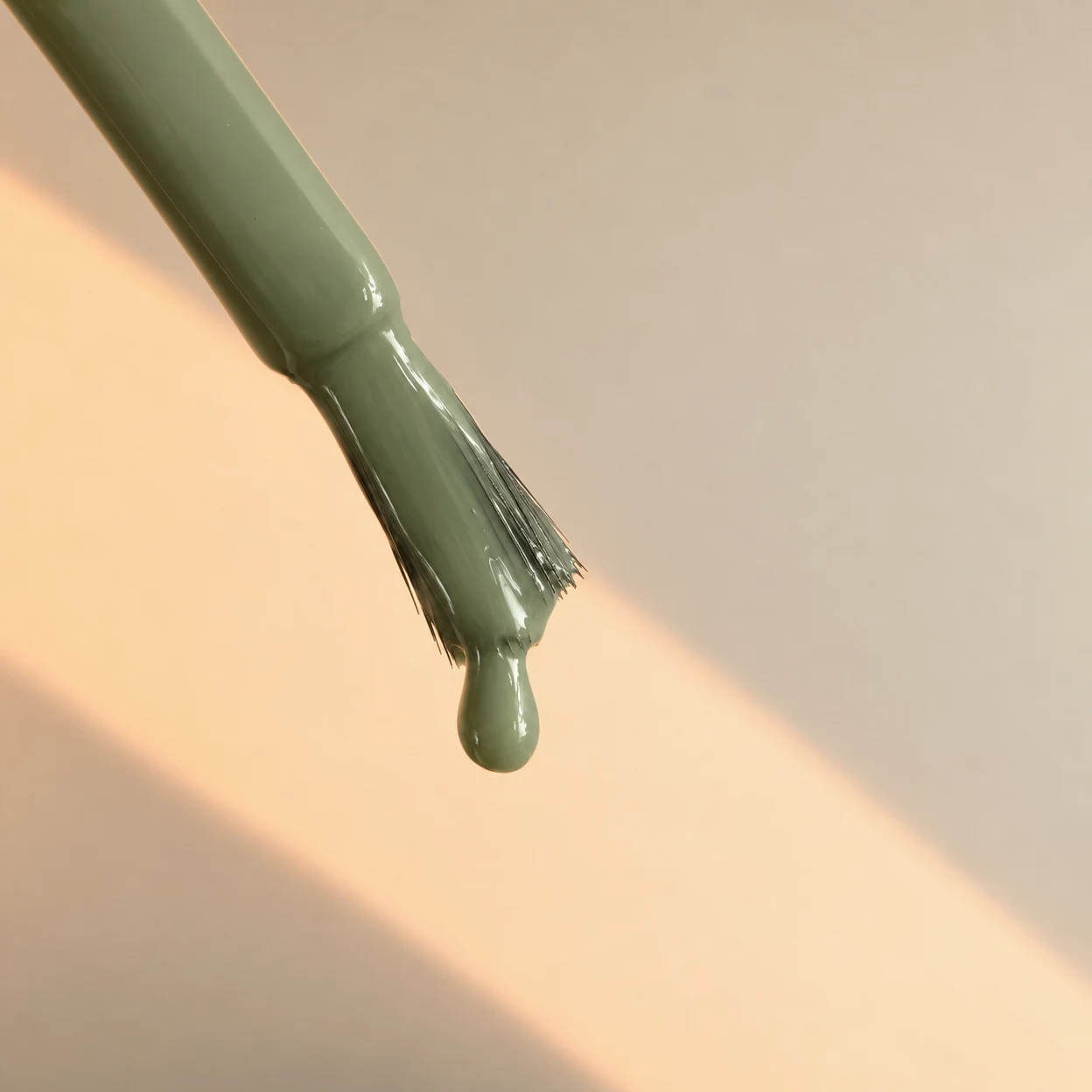 An olive-sage nail-polish brush enters diagonally from the upper-left, its lacquer-coated bristles holding a rounded drop against a cream backdrop crossed by a…