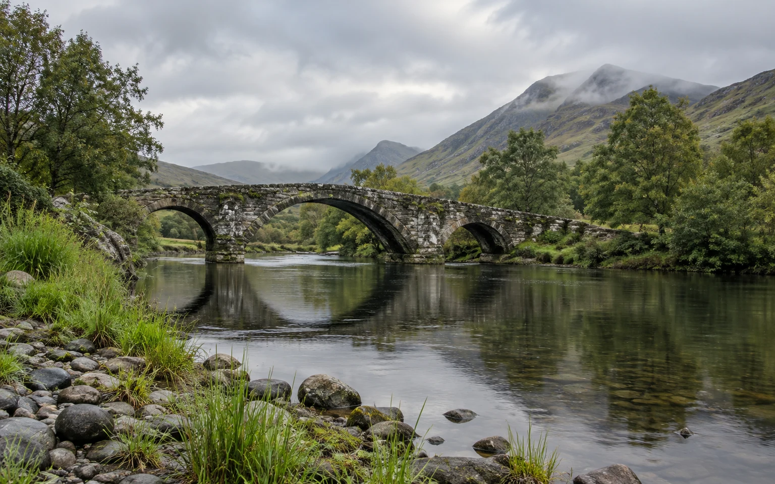 A weathered three-arch stone bridge spans a calm river in a green valley, with reeds and rounded stones along the foreground bank, trees at the water’s edge…