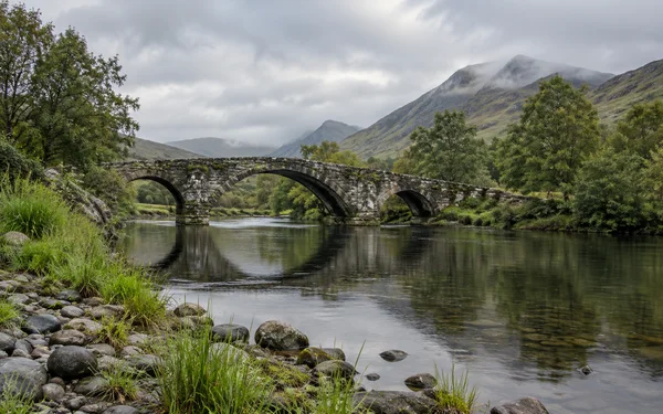 A weathered three-arch stone bridge spans a calm river in a green valley, with reeds and rounded stones along the foreground bank, trees at the water’s edge…