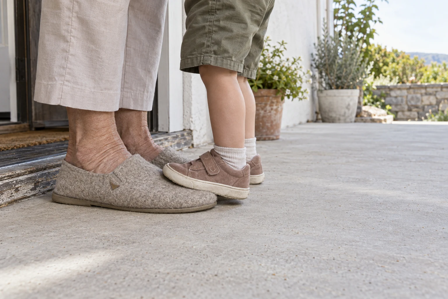 At an open patio doorway, a child in muted clay Velcro sneakers and short socks stands beside an older adult in cropped light trousers and oat-colored felt…