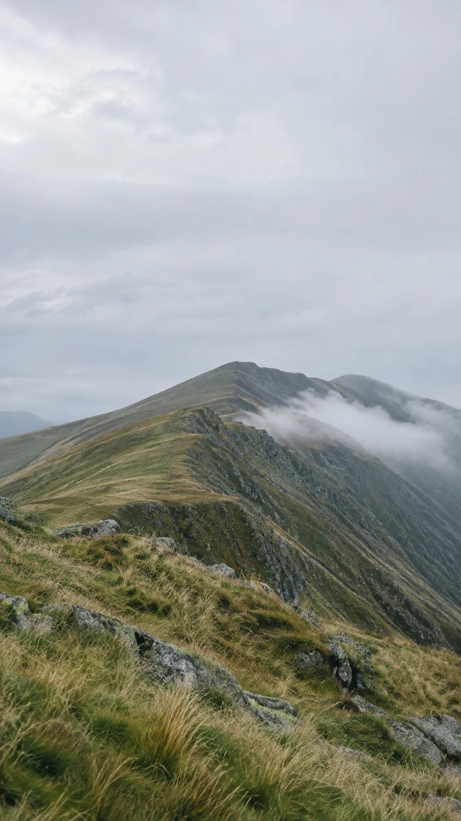 A grassy, rocky mountain ridge runs diagonally toward a higher crest beneath a broad pale grey sky, with a soft band of cloud drifting along the far slope.