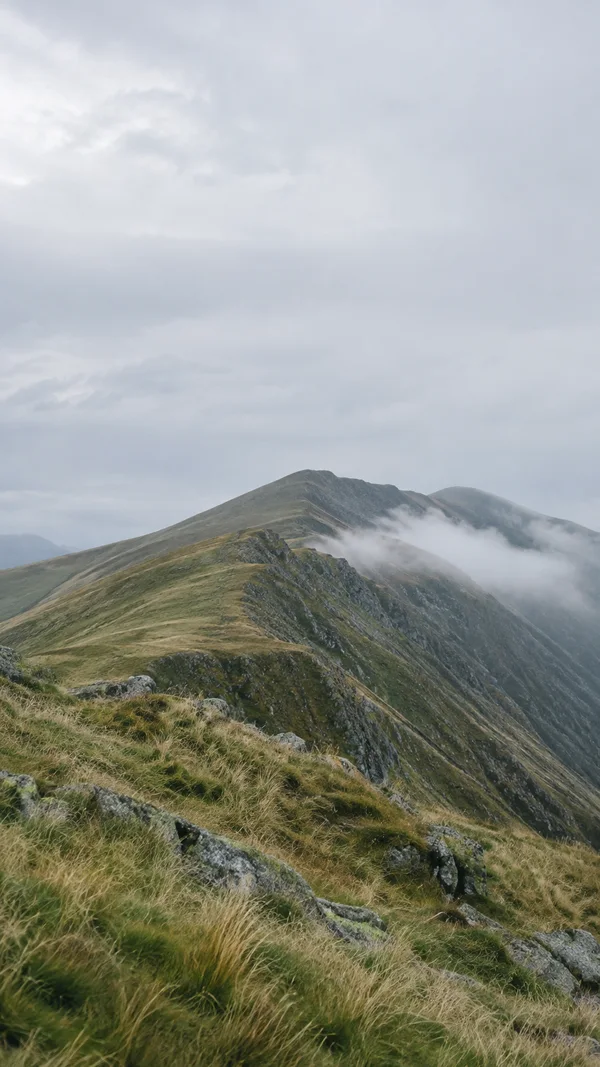 A grassy, rocky mountain ridge runs diagonally toward a higher crest beneath a broad pale grey sky, with a soft band of cloud drifting along the far slope.