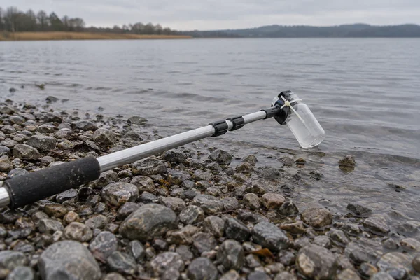 A telescoping metal pole with a clear plastic bottle attached lies diagonally across wet shoreline pebbles, with the bottle extending over a calm grey lake; a…