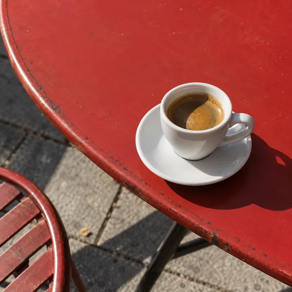 A small white espresso cup and saucer sit near the curved edge of a worn red metal café table, with grey pavement and part of an empty red chair visible below.
