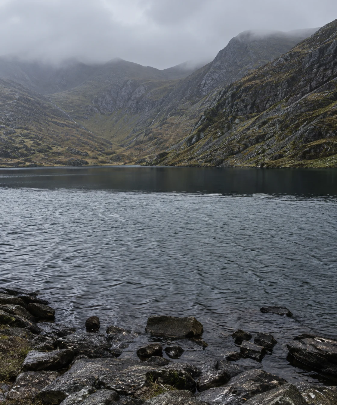 A mountain lake sits beneath low cloud, with steep rocky slopes surrounding a broad, dark band of still water above wind-rippled water and wet shoreline stones.