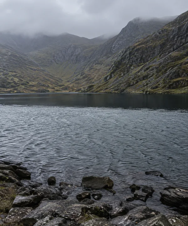 A mountain lake sits beneath low cloud, with steep rocky slopes surrounding a broad, dark band of still water above wind-rippled water and wet shoreline stones.