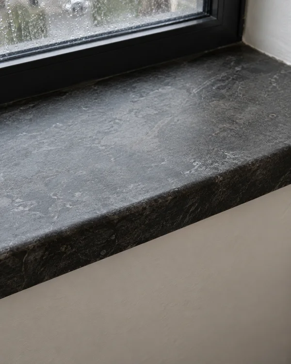 A dark charcoal stone windowsill with a rounded, worn front edge beneath a rain-speckled window, viewed diagonally above a pale plaster wall.