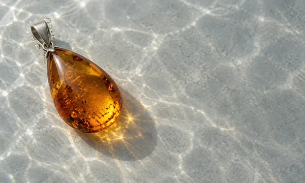 A large polished amber teardrop pendant with a silver-toned bail rests on a pale grey stone surface, surrounded by bright rippling light patterns and small…