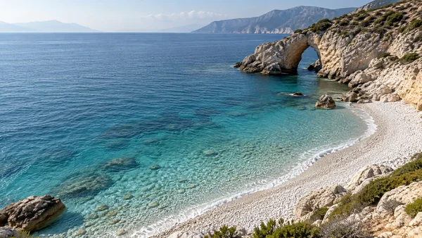 Elevated view over a white-pebble cove with clear blue water, a pale rocky headland and a sea arch; distant mountains line the horizon beneath a hazy sky.