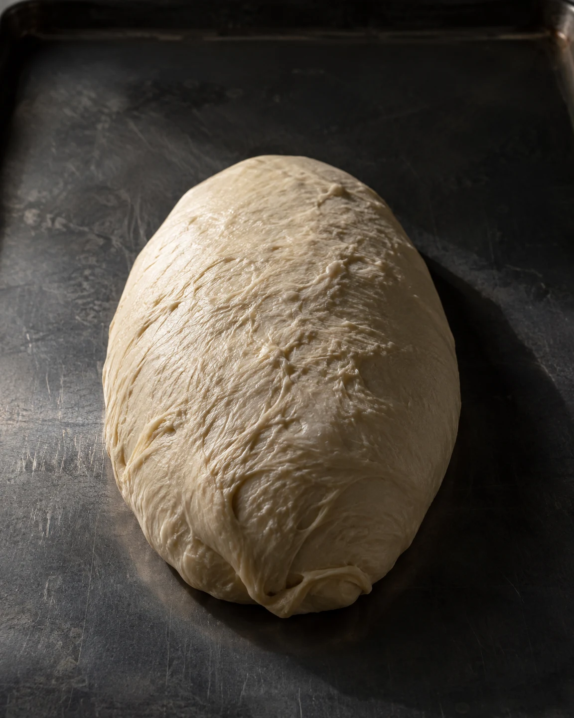 A pale oval of uncooked dough with an irregular folded seam rests lengthwise on a dark steel baking sheet, lit from the upper left and casting a shadow to the…
