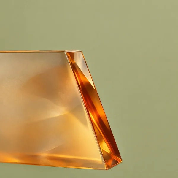 A large translucent amber glass form fills the left side of a square image, with a vivid russet-orange diagonal bevel against a pale olive background and open…