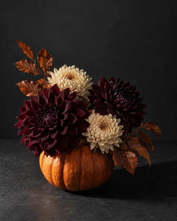 A pumpkin-shaped orange vessel holds two deep burgundy flowers, two cream flowers and copper-brown leaves on a charcoal tabletop against a dark charcoal…