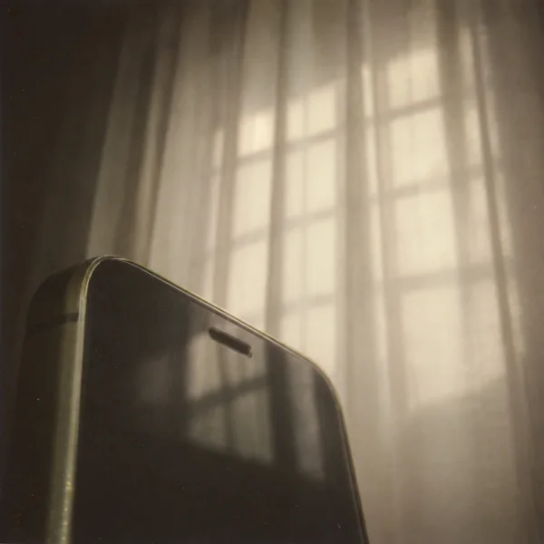 An extreme close-up of a dark-screened phone with a muted olive edge fills the lower-left of the frame; warm cream curtains behind it show soft rectangular…